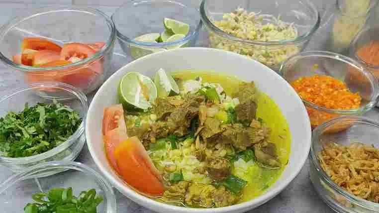 Semangkuk soto daging sapi dengan kuah gurih kuning, disajikan bersama tomat, jeruk nipis, tauge, sambal, bawang goreng, dan daun seledri.