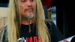 Seorang pria berambut panjang pirang dengan jenggot tipis sedang memegang selembar kertas sambil mengenakan kaus hitam bergambar.