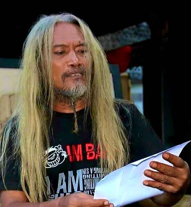 Seorang pria berambut panjang pirang dengan jenggot tipis sedang memegang selembar kertas sambil mengenakan kaus hitam bergambar.