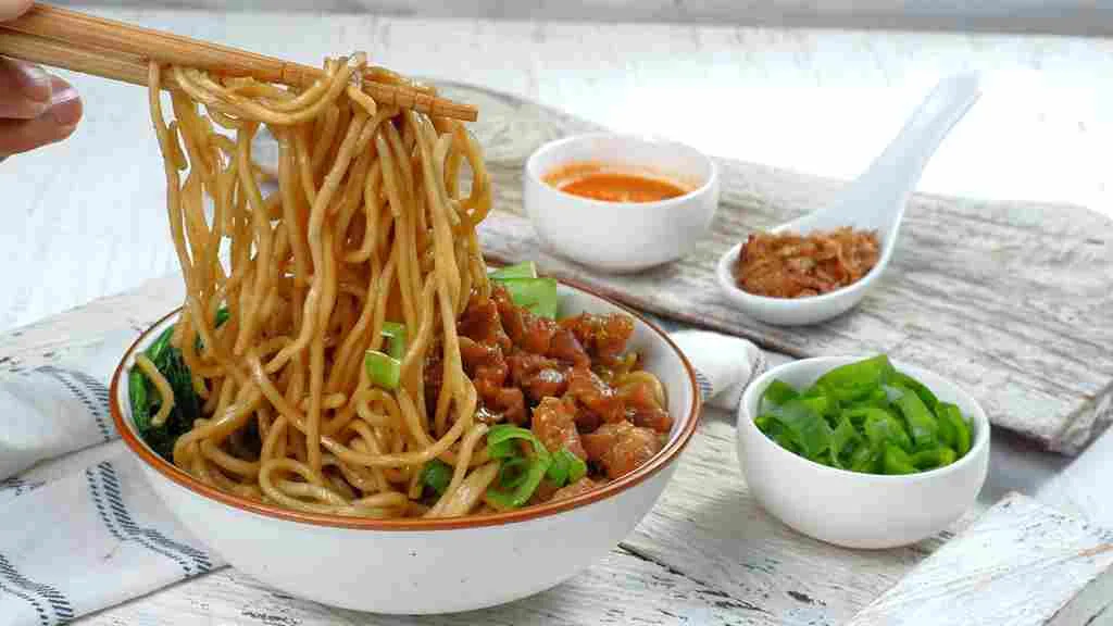 Mie ayam enak dengan topping ayam kecap, sawi hijau, dan taburan bawang goreng, disajikan dengan kuah dan sambal pelengkap.