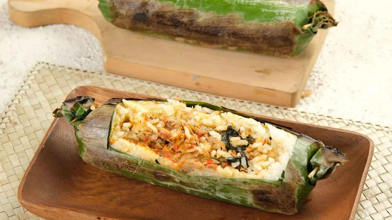 Nasi bakar ayam kemangi dalam balutan daun pisang, disajikan di atas piring kayu dengan aroma gurih dan harum khas bakaran.