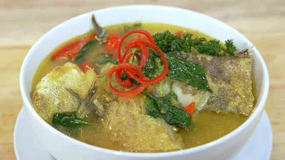 Semangkuk ikan kuah kuning dengan kuah berwarna kuning cerah, potongan ikan kerapu, daun kemangi, dan irisan cabai merah sebagai hiasan.