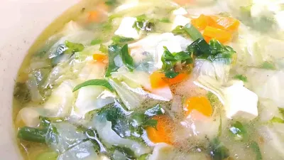 Semangkuk sayur sop bening berisi wortel, kubis, tahu, daun bawang, dan seledri yang tampak segar dan menggugah selera.