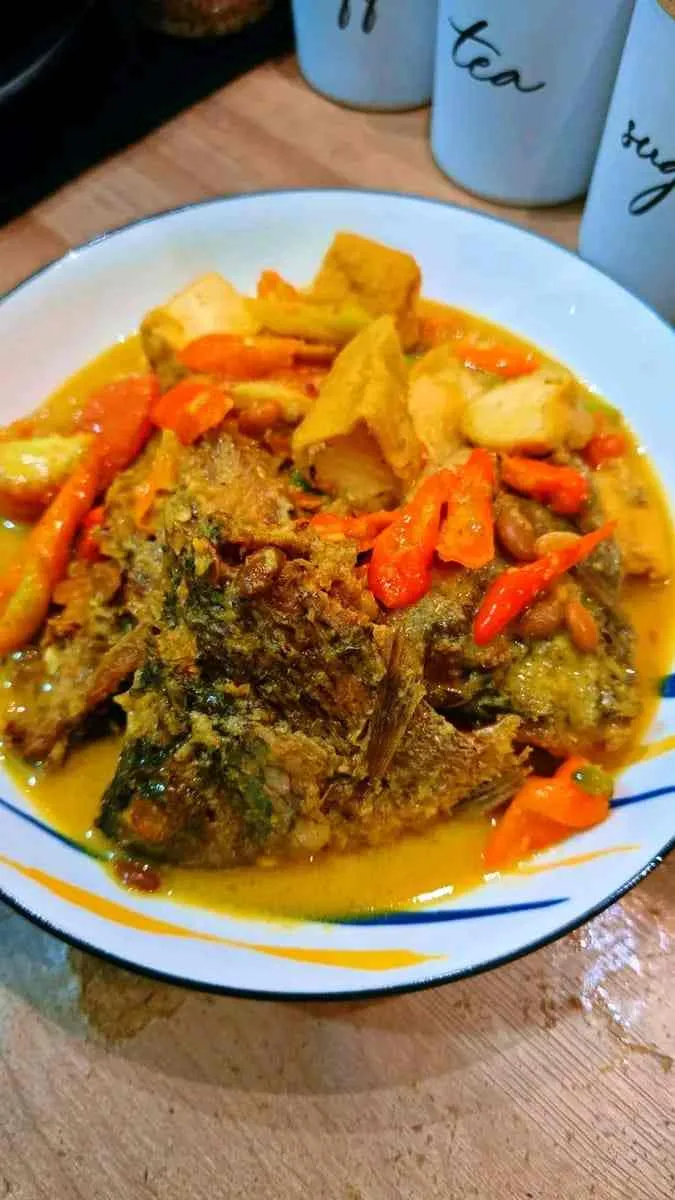 Ikan nila tauco tahu dengan kuah kuning gurih dan potongan cabai merah disajikan di piring putih di atas meja kayu.