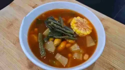 Semangkuk sayur asem berkuah bening kemerahan berisi jagung, kacang panjang, melinjo, dan potongan labu siam di atas talenan kayu.
