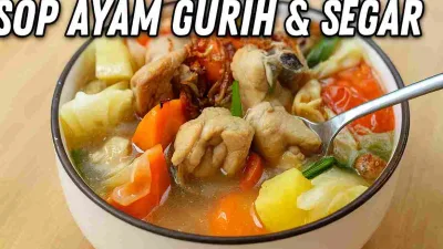 Semangkuk sop ayam rumahan berisi potongan ayam, wortel, kentang, kol, dan tomat dengan kuah bening gurih, diambil dekat dengan sendok yang sedang menyendok potongan ayam.