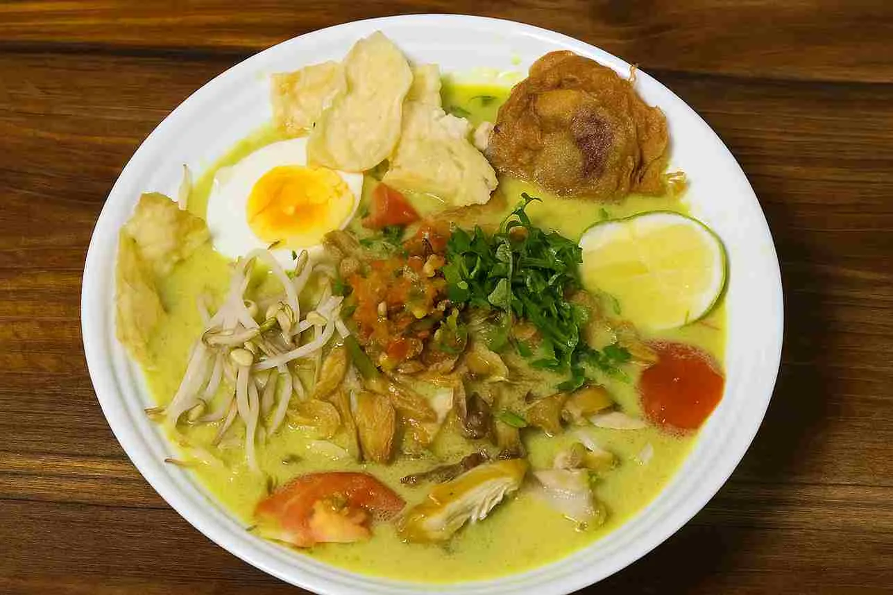 Semangkuk Soto Ayam Medan lengkap dengan telur rebus, perkedel, tauge, sambal, dan irisan jeruk nipis disajikan di atas piring putih dengan kuah kuning santan gurih di atas meja kayu.