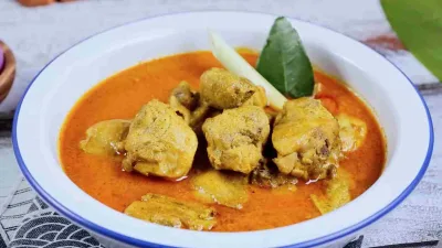 Kari ayam kuah santan berwarna kuning kemerahan dalam mangkuk putih, disajikan dengan potongan ayam, serai, dan daun jeruk.