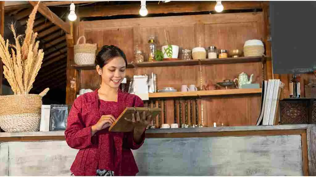 Perempuan mengenakan kebaya merah berdiri di kafe bernuansa tradisional sambil memegang tablet, memadukan budaya dan gaya modern.